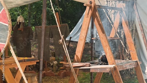 A medieval reenactment area with wooden frame structures and smoke rising from a fire. Individuals dressed in simple cloaks engage in various activities, reflecting life in the early Middle Ages