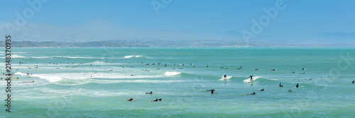 Wide panorama of surfers paddling out and waiting for rolling Atlantic waves off Plage de la Côte des Basques under a clear sky in Biarritz France