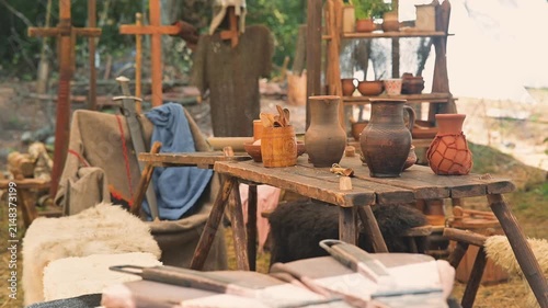 A medieval setting shows a wooden table with pottery and tools arranged neatly. Various handmade items are visible alongside simple fabrics and natural surroundings