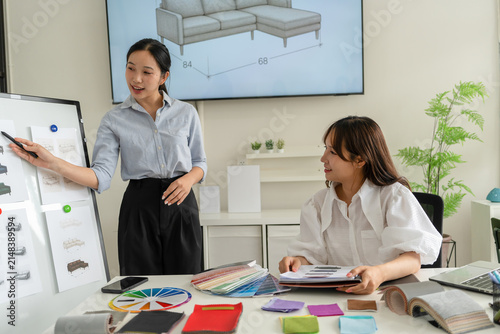 Two Asian women working together in a bright, modern studio.while using a laptop and smartphone. The creative workspace is filled with colorful samples for seasonal palette selection.