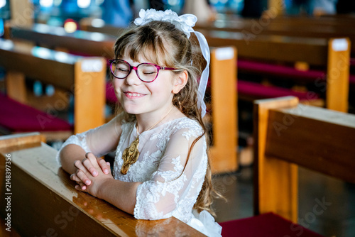 Canvas Print Child girl in traditional white first communion dress kneeling in prayer with folded hands inside a Catholic church during a religious celebration