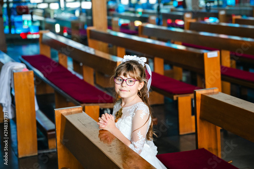 Photography Child girl in traditional white first communion dress kneeling in prayer with folded hands inside a Catholic church during a religious celebration