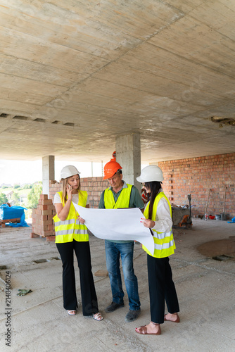 Engineer team in vests studies blueprint near columns. Supervisor points at drawing and explains sequence assigning task before work starts