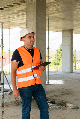Construction builder in safety vest thinks about work process. Experienced constructor touches face with hand and inspects building analyzing results