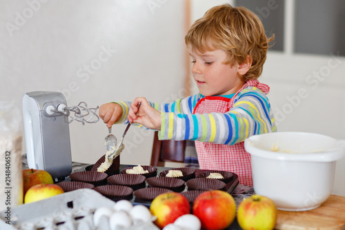 Cute little happy blond preschool kid boy baking apple cake and muffins in domestic kitchen. Funny lovely healthy child having fun with working with mixer, flour, eggs, fruits. Little helper indoors