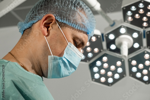 Man surgeon keeps side profile under bright surgical lamps in operating room. Focused medic looks down preparing precise sterile action
