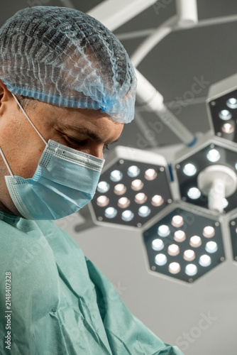 Man surgeon bends under surgical lamps in operating room. Focused male medic lowers gaze as prepares sterile work with calm precision