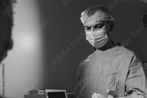 Man surgeon stands in dim operating room during sterile procedure. Focused medic lowers gaze watching gloved hands with calm precision