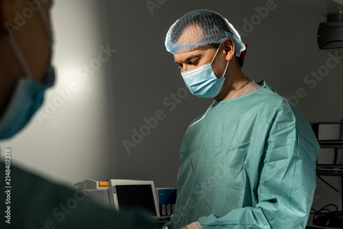 Man surgeon lowers gaze during sterile procedure in operating room. Focused medic studies surgical field preparing next careful step