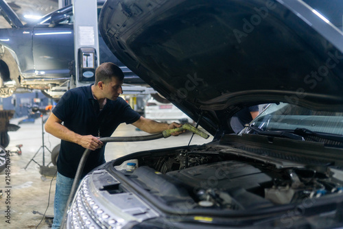 Mechanic cleans car engine with vacuum cleaner before diagnostics in shop. Man worker hoovers vehicle under hood area at service station
