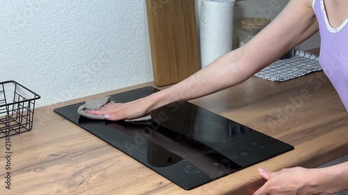 Close up of woman's hands wiping a modern black two-burner induction cooktop embedded in a wooden kitchen countertop. Cleaning and domestic chores concept.