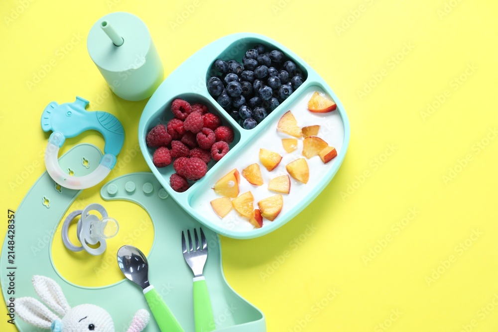 Obraz premium Baby tableware. Section plate with food, bottle, cutlery, silicone bib, toys and pacifier on yellow background, flat lay. Space for text