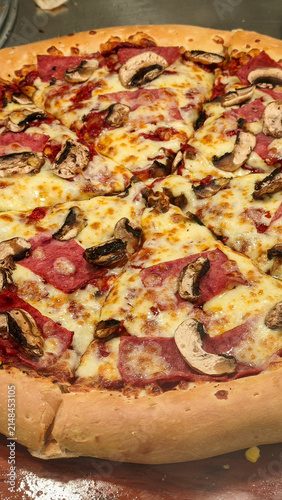 Freshly baked gourmet pizza with salami, mushrooms, and melted mozzarella, sold in a glass display case. 