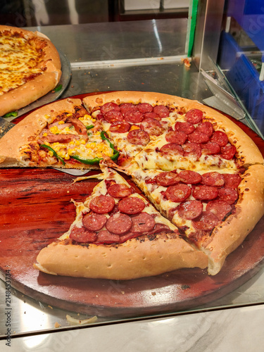 Freshly baked slices of pepperoni and Supreme pizza are sold from a glass display case. A mouthwatering  pizza featuring rich melted cheese, savory pepperoni slices, sweet corn, and green peppers.