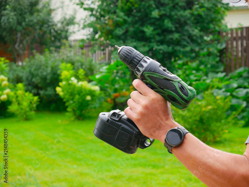 Bild auf Leinwand Close-up of a man hand wearing a black watch holding a green and black cordless drill against a blurred green lawn and garden background