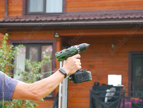 Arm and hand wearing a smartwatch holding a green cordless drill in front of a wooden cabin wall with a window.