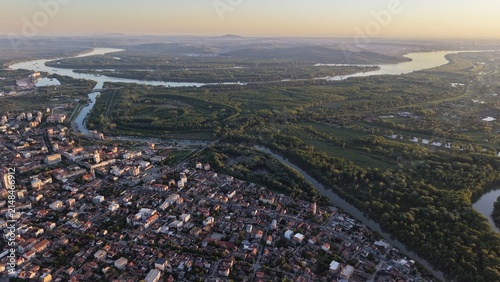 Aerial view of the confluence of the Tamis and Danube rivers, featuring urban rooftops, lush riverine forests, and golden hour light in Pancevo, Serbia.