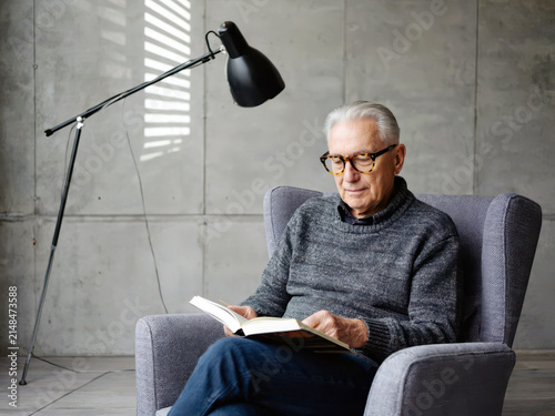 Older white man in grey sweater and glasses seated in light grey armchair reading book under black arc lamp in clean concrete interior. Calm reading concept