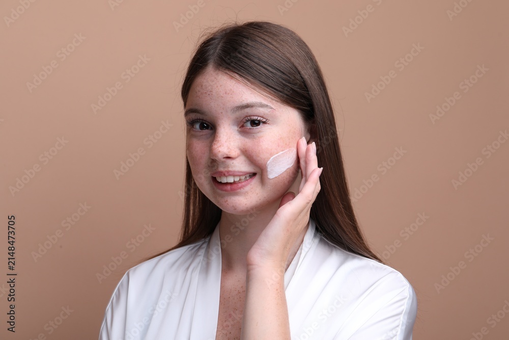 Obraz premium Beautiful girl with freckles applying cream on her face against dark beige background