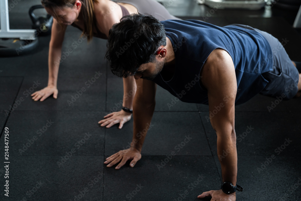 Fototapeta premium Young Asian couple holds plank position during bodyweight floor workout in modern gym, man and woman building core strength, endurance, motivation, discipline, healthy teamwork lifestyle, balance.