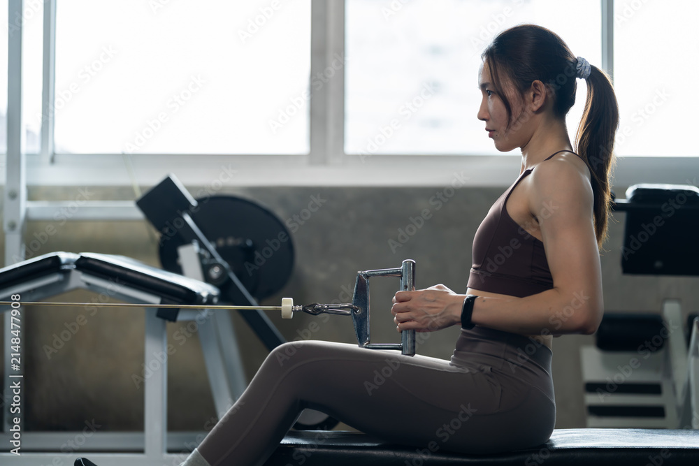 Fototapeta premium Young Asian woman pulling cable row handle toward torso in modern gym, seated strength training for back muscle, healthy lifestyle, fitness routine, wellness, determination, workout motivation concept