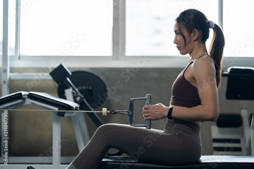 Young Asian woman pulling cable row handle toward torso in modern gym, seated strength training for back muscle, healthy lifestyle, fitness routine, wellness, determination, workout motivation concept