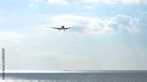 Commercial airliner flying on landing approach low over the sparkling sea under a blue sky with clouds in bright sunlight, wide angle static shot