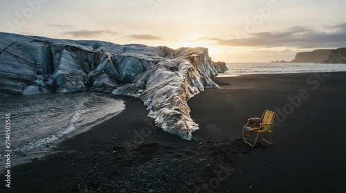 Wooden chair on a black sand beach beside a glacier under a cloudy sunset