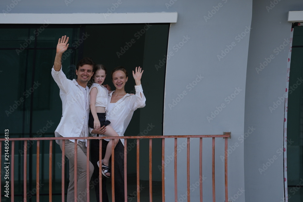 Obraz premium Caucasian family of three embracing wave happily from their balcony, the girl riding piggyback on the balcony of their new house overlooking the residential estate, authentic everyday scene