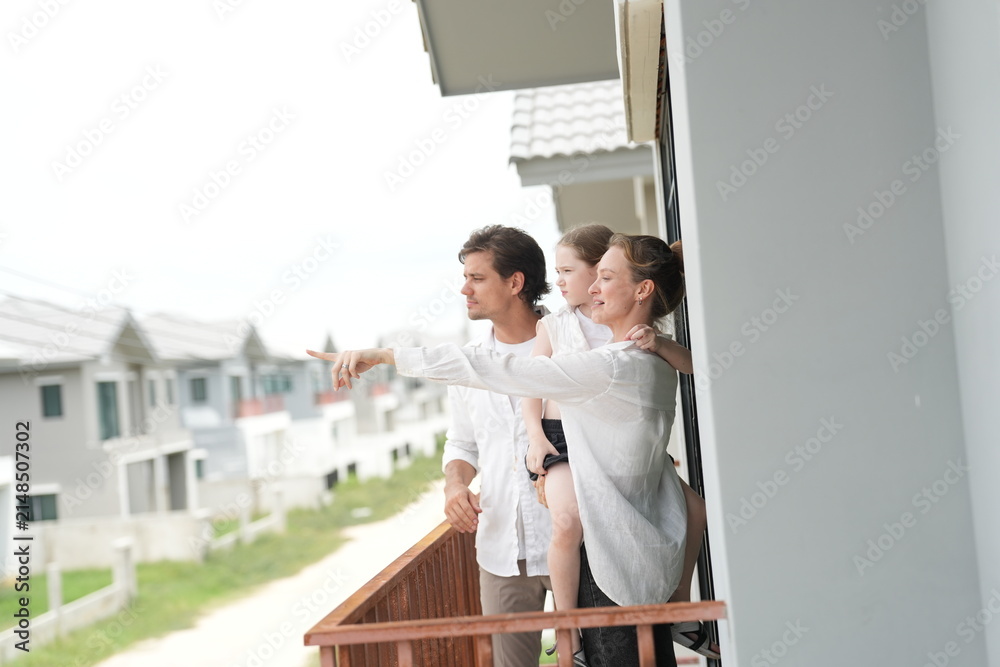 Obraz premium Happy young family with little daughter enjoy the view of the growing suburb from their balcony on the upper balcony of their new home above the neighborhood, candid real moment