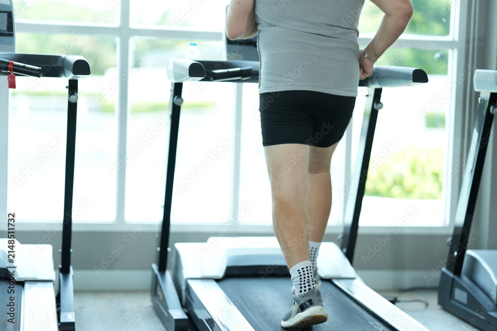Fototapeta premium Funny fat senior man in sportswear walks the treadmill in close up, step after heavy step at the start of his new healthy lifestyle in the gym on a bright sunny day