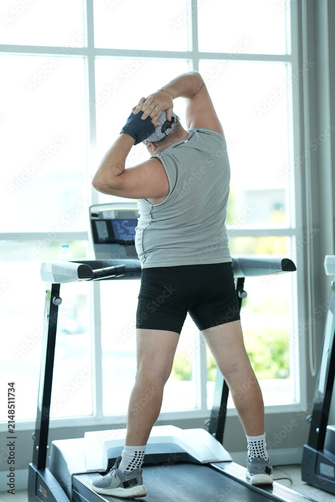 Fototapeta premium Funny fat senior man in sportswear does standing side stretches at the bright window front in a bright modern gym with large windows. Humorous diet motivation and fitness struggle concept