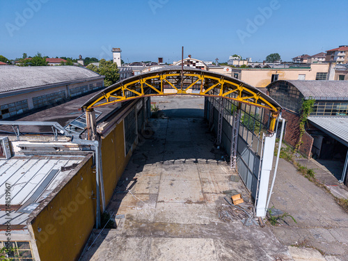 Aerial view of an abandoned industrial warehouse with a yellow metal arch, weathered concrete flooring, and old factory buildings in Casale Monferrato, Piedmont, Italy.