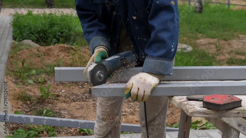Gloved worker cutting gray metal beam outdoors with handheld grinder, sending bright sparks across rough wooden bench, worn workwear, grassy yard, and muted daylight, with construction, worker