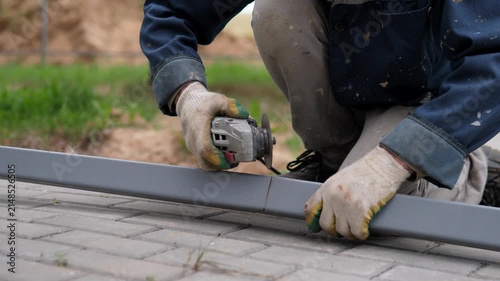 Gloved worker kneels beside gray metal bar, using handheld grinder outdoors in daylight and sending bright sparks across paved ground during repair work, with worker, grinding, cutting, spark detail