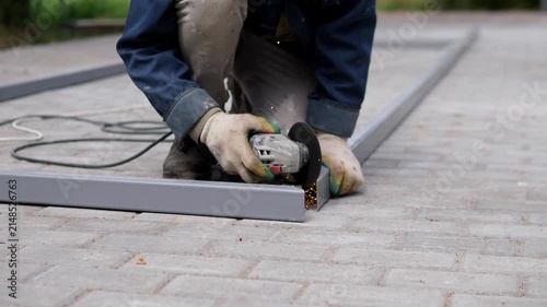 Worker kneels on paved ground and uses handheld grinder on gray metal edging, creating bright sparks beside blurred outdoor greenery and rough stone surface, with worker, kneeling, grinding, spark