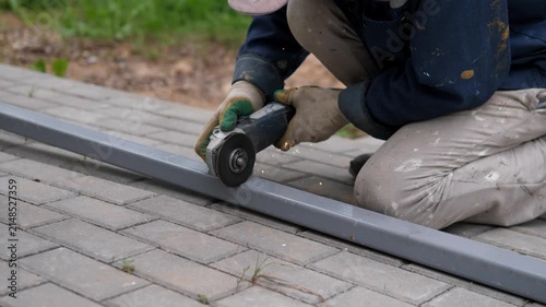 Construction worker kneels beside paved walkway while holding handheld grinder against long gray metal rail, with visible sparks, gloved hands, blurred greenery, muted tones, and practical