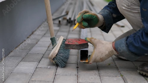 Crouched worker in protective gloves holds yellow pencil against metal tube beside brush and broom on gray pavers, suggesting careful measuring or fitting during outdoor repair work with soft