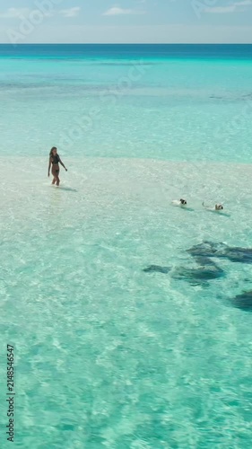 Breathtaking aerial view of a young woman enjoying a leisurely walk on a pristine sandbank, her friend swimming in the crystal-clear turquoise waters of the maldives