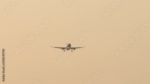 Commercial passenger airplane descending head-on with landing gear down and headlights on, flying through warm golden sunset sky, telephoto shot