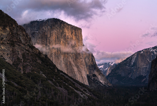 View of the towering El Capitan granite monolith rising above the valley floor with dense pine forests and soft pink sunset light in Yosemite National Park, California, United States.