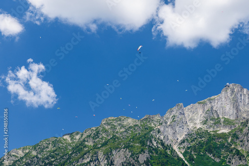 View of paragliders soaring over the rugged peaks of the Mont Blanc massif under a bright blue sky with scattered clouds in Chamonix, Auvergne-Rhone-Alpes, France.
