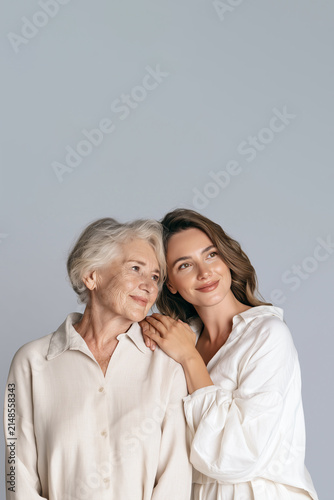 Elderly Woman and Younger Woman Standing Together in White Shirts against a plain light gray background, Showing Generational Bond, Family Love and Tender Support with Copy Space, Vertical Aspect 2:3