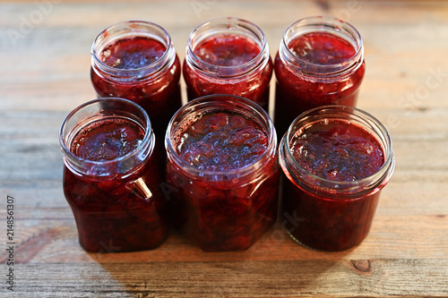 Six open glass jars filled with homemade red berry jam stand on a rustic wooden table
