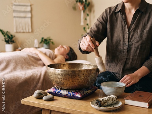 Professional spa therapist performing a relaxing sound healing session with a metal singing bowl for a client lying on a massage table in a studio