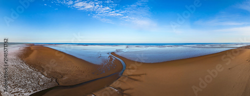Aerial view captures coastal salt flats and winding river. Cracked white earth meets green grassland. Sandy path curves beside waterway. Friskney Flats, Lincolnshire UK, Skegness