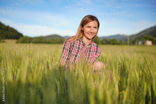 Young woman wearing a shirt crouching in unripe green wheat field, smiling