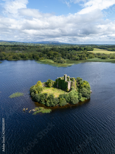Aerial view of McDermott Castle situated on a small island in the middle of a lake, surrounded by lush green trees and calm dark water in County Roscommon, Ireland.