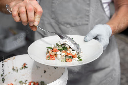 Male chef in gray apron plating fresh vegetable salad with spoon from a white bowl onto a round plate in a bright kitchen setting