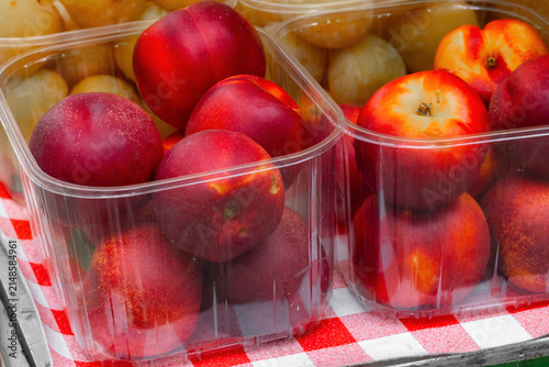 Fresh red nectarines and yellow plums displayed in clear plastic containers on a checkered tablecloth at a local market